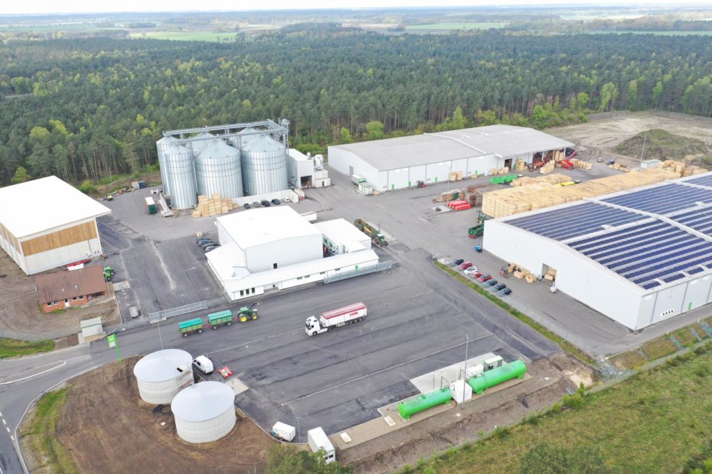 Luftaufnahme eines Industriegebiets mit Silos, Lagerräumen und Transportfahrzeugen in einer ländlichen Umgebung.