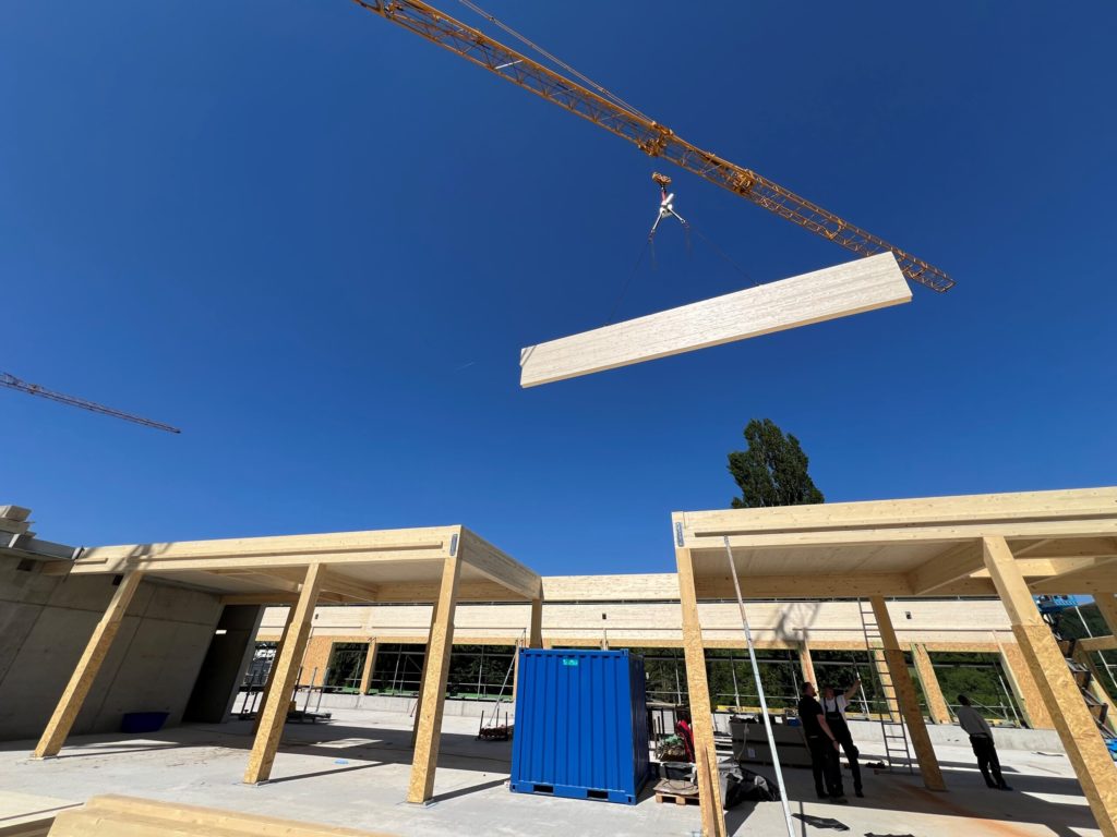 Ein Kran hebt ein Holzbauelement auf einer Baustelle unter klarem Himmel mit Bauarbeitern und Materialien im Hintergrund.