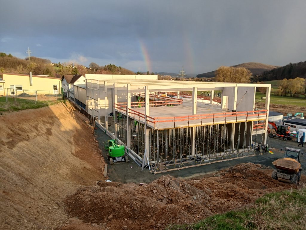 Bau eines neuen Gebäudes mit sichtbarer Baustelle, Gerüst und Maschinen, umgeben von hügeliger Landschaft und Regenbogen.
