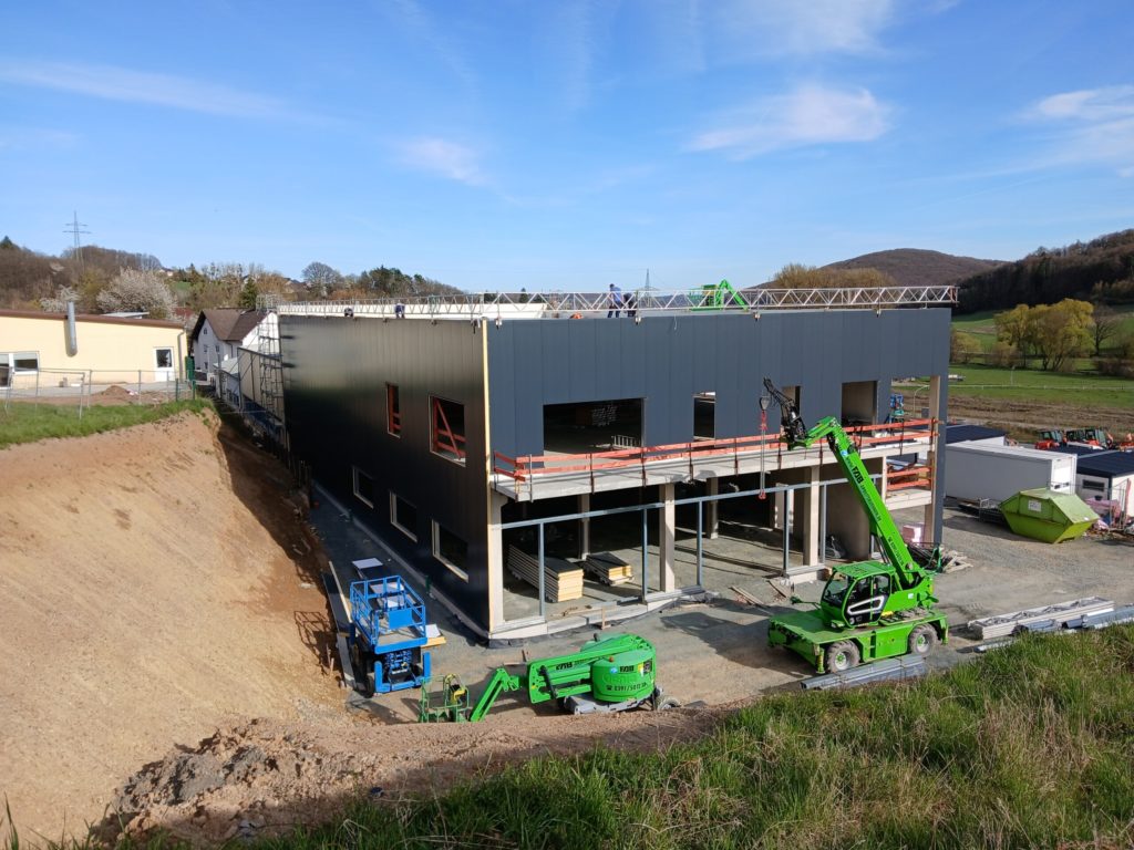 Baufortschritt der Erweiterung einer Lagerhalle mit Büroräumen, inklusive Baugeräten und Arbeiter auf dem Dach.
