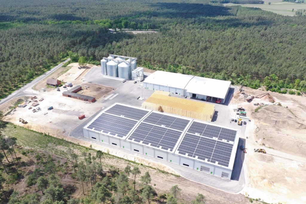 Luftaufnahme eines modernen Produktionsstandorts mit Solaranlagen und silos, umgeben von Wald.