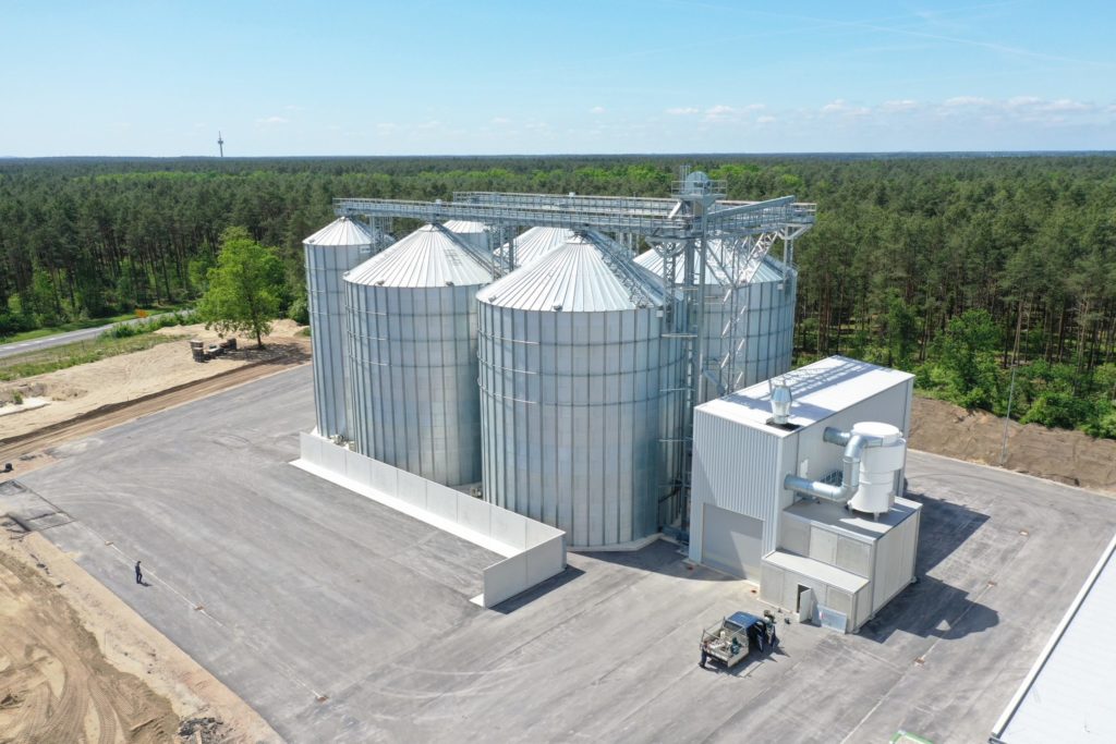Moderne Silos und Anlagen zur Lagerung von Getreide in ländlicher Umgebung mit Wald im Hintergrund.