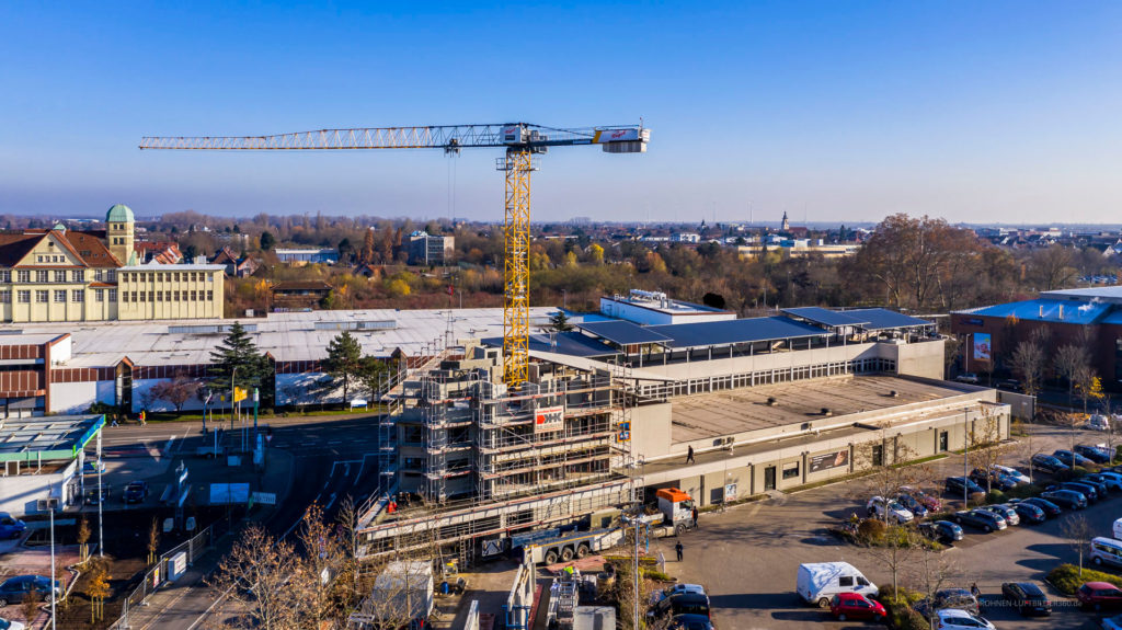 Bauarbeiten an einem modernen Gebäude mit einem großen Kran, umgeben von Parkplätzen und einer städtischen Landschaft.