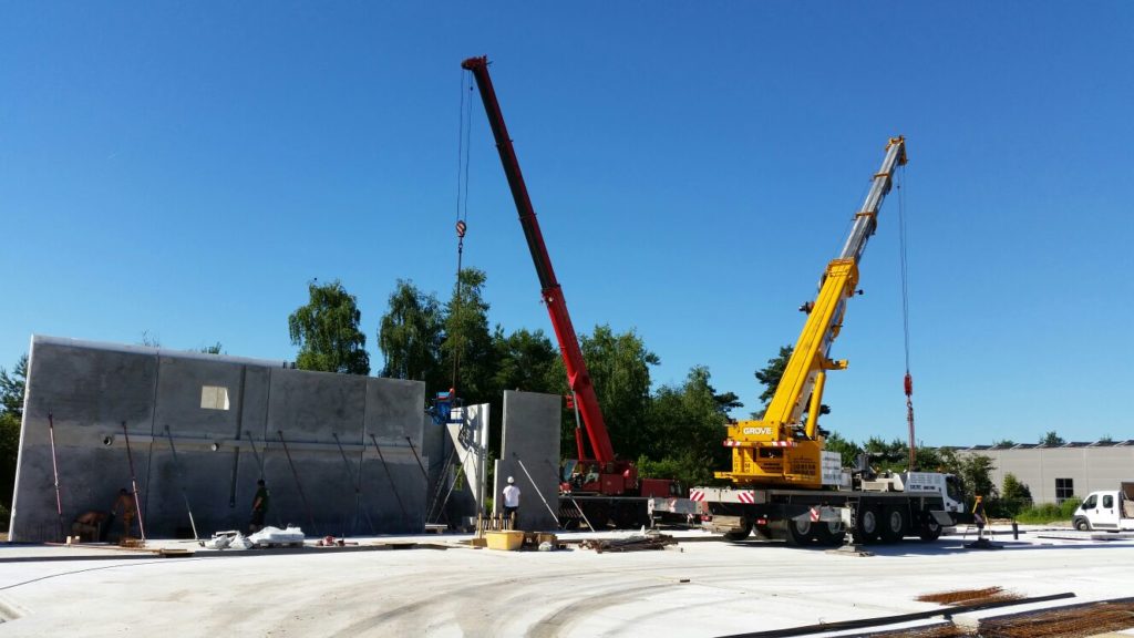 Bauarbeiten mit zwei Kränen, die Betonwände heben, unter blauem Himmel auf einer Baustelle.