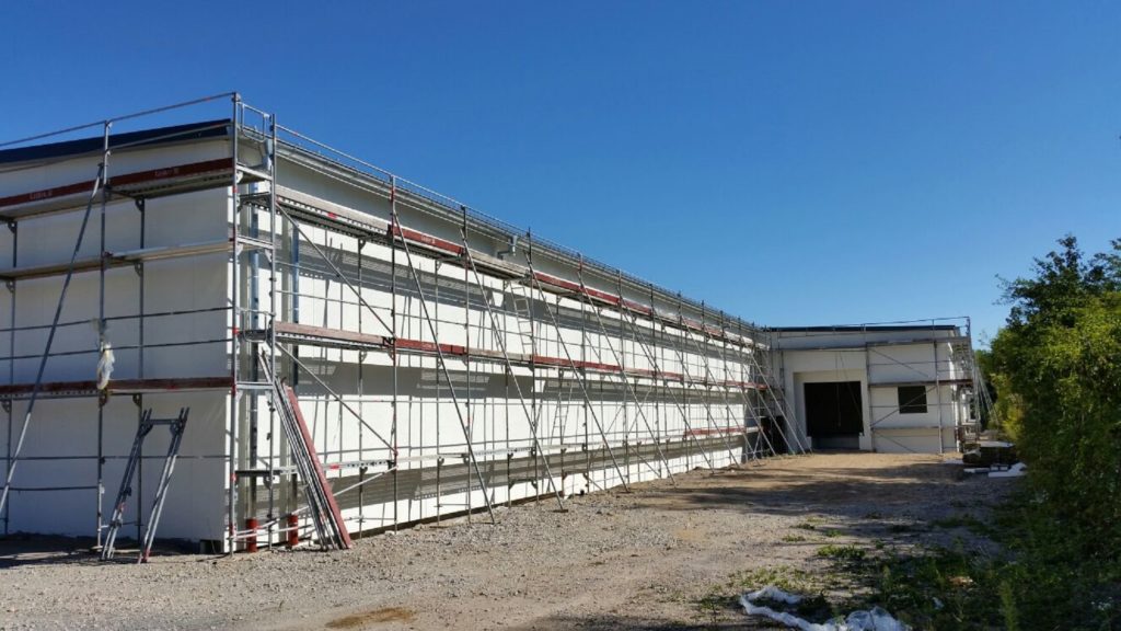Eine Baustelle mit einem Gebäude, das mit Gerüsten für Malerarbeiten umgeben ist, unter klarem blauen Himmel.