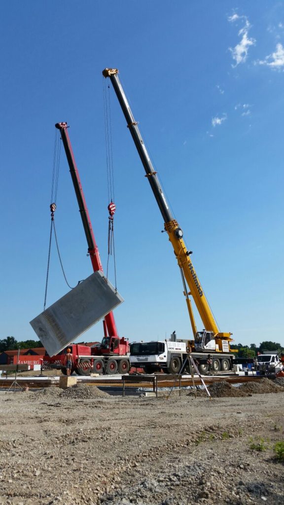 Zwei Kräne heben ein Betonstück auf einer Baustelle unter blauem Himmel.