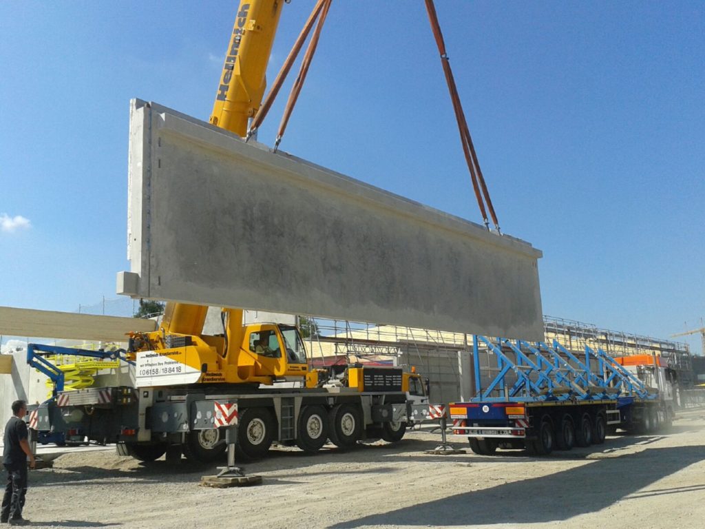 Ein Kran hebt ein großes Betonbauteil auf einer Baustelle, während Arbeiter und Maschinen im Hintergrund sichtbar sind.