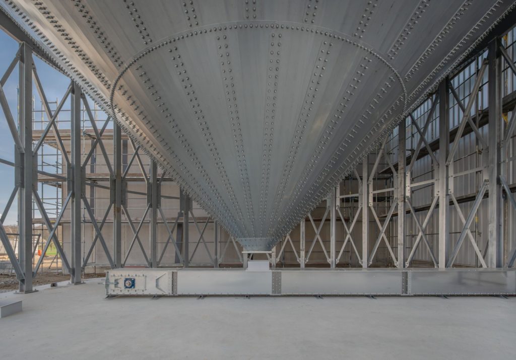 Blick auf eine große, metallische Trichterstruktur in einem Bauwerk mit sichtbaren Stahlrahmen und Baustellenumgebung.