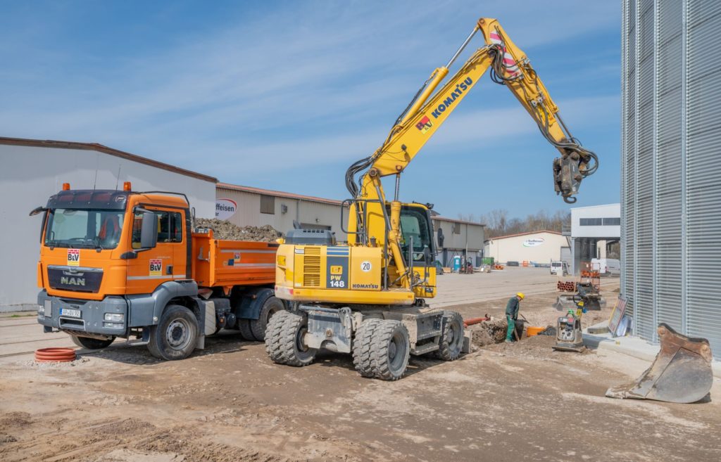 Bauarbeiten mit einem gelben Komatsu-Bagger und einem orangefarbenen MAN-Lkw auf einer Baustelle bei klarem Himmel.