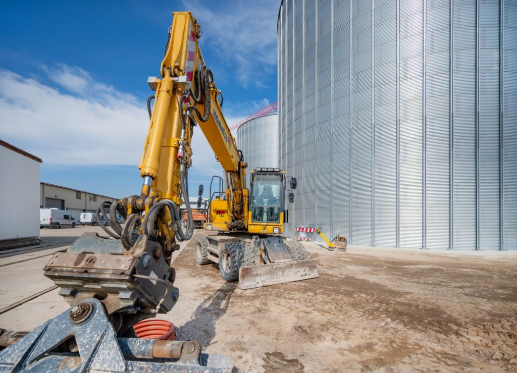 Bagger arbeitet auf einer Baustelle neben großen Silos unter klarem Himmel.