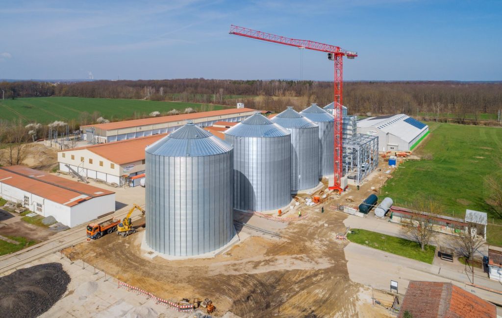 Neue Silos auf einem landwirtschaftlichen Gelände, ergänzt durch einen Kran im Hintergrund, unter blauem Himmel.