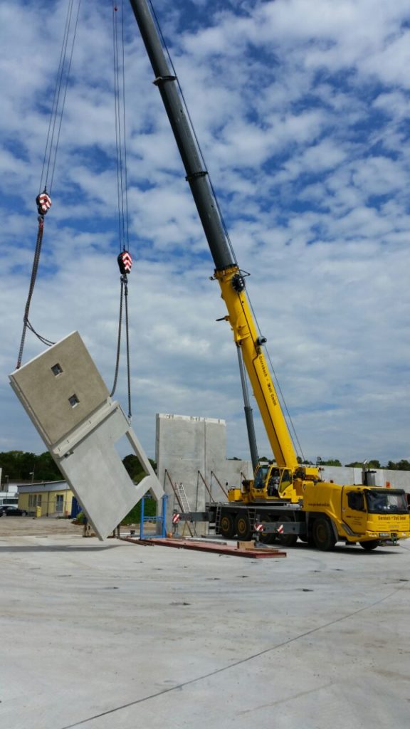 Kran hebt eine Betonwand auf einer Baustelle unter einem bewölkten Himmel.
