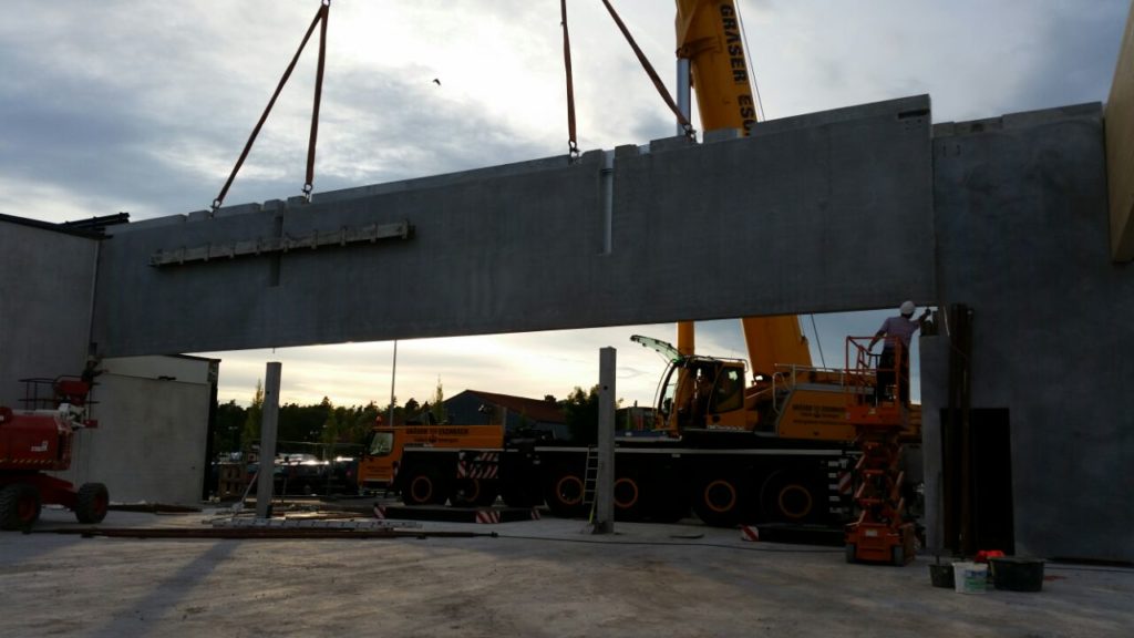 Baustelle mit Kran, der eine große Betonplatte hebt, während Arbeiter an der Montage eines Gebäudes beteiligt sind.