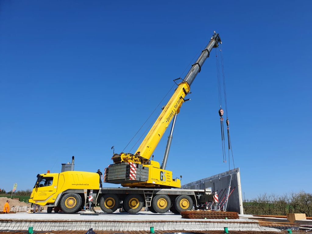 Ein gelber Kran hebt Bauplatten auf der Baustelle eines neuen ALDI-Marktes in Schweighofen unter klarem blauen Himmel.