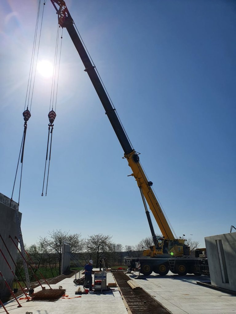 Ein Kran hebt Materialien auf einer Baustelle unter klarem Himmel, während Arbeiter im Vordergrund tätig sind.