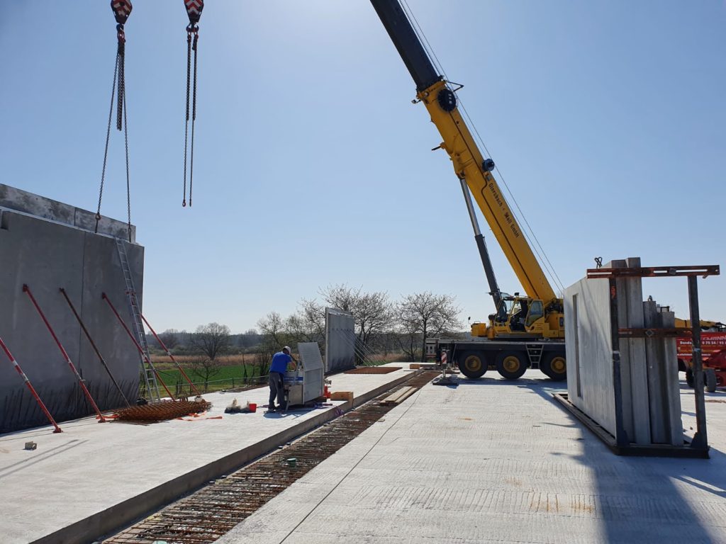 Bauarbeiter und Kran heben Betonwände auf einer Baustelle bei klarem Himmel und frühlingshaftem Wetter.