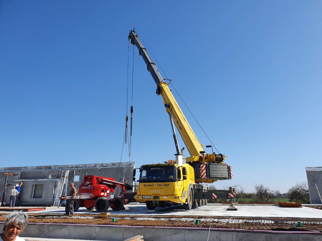 Ein gelber Kran hebt Bauelemente auf einer Baustelle, während Arbeiter und ein roter Arbeitsbühnenlift im Hintergrund sichtbar sind.