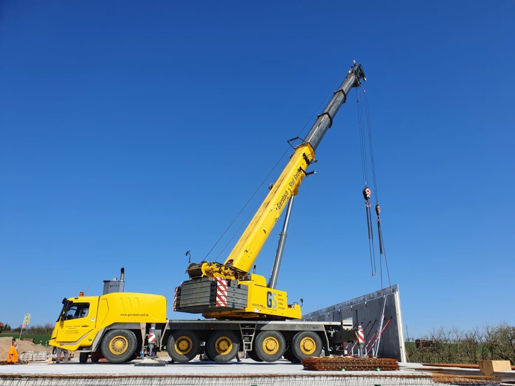 Ein gelber Kran hebt eine Wand auf einer Baustelle unter klarem Himmel.