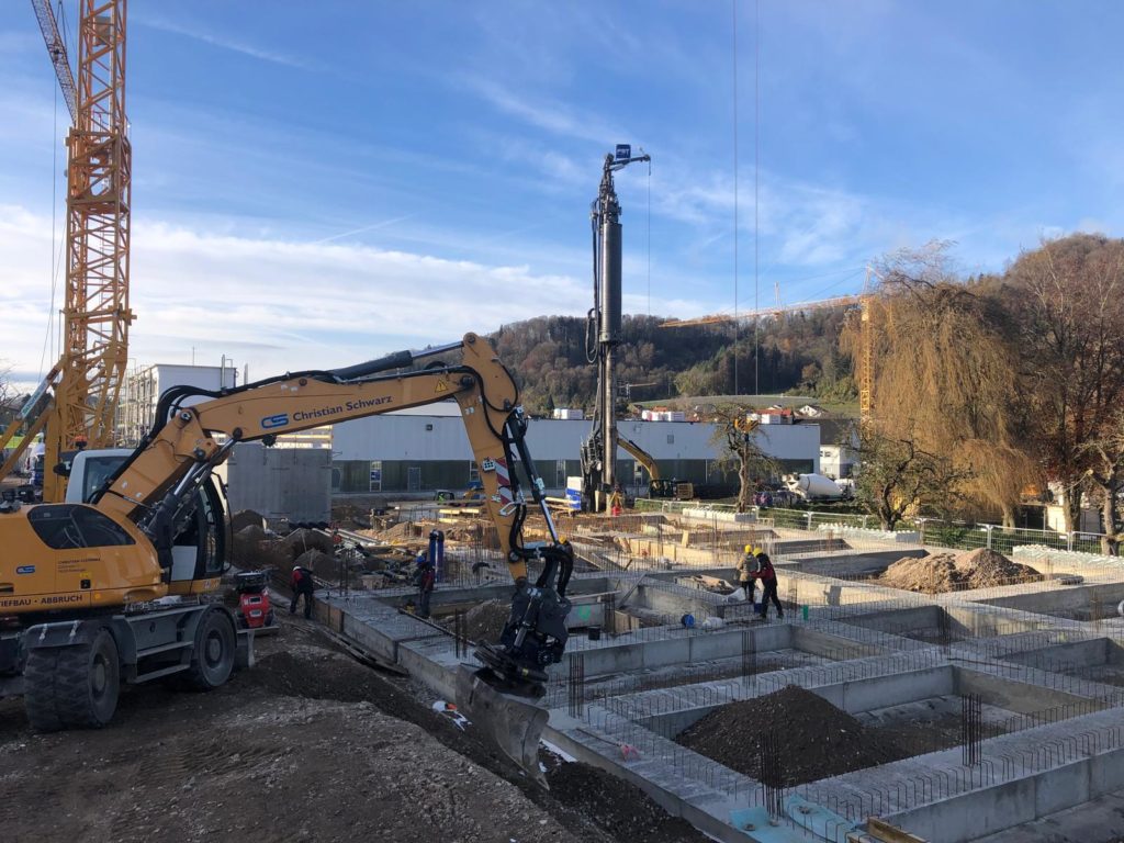 Bauarbeiten auf einer Baustelle mit Bagger, Bohrmaschine und mehreren Arbeitern unter blauem Himmel.