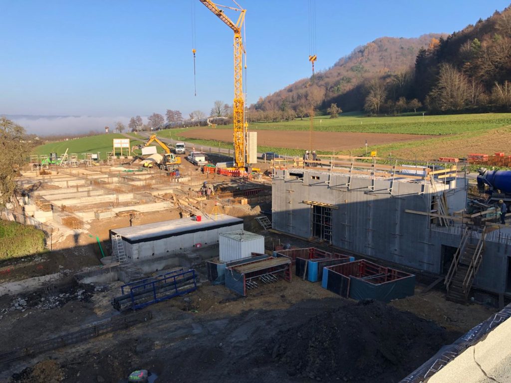Bauplatz mit Kränen, Betonfundamenten und Baustellenfahrzeugen in ländlicher Umgebung unter klarem Himmel.