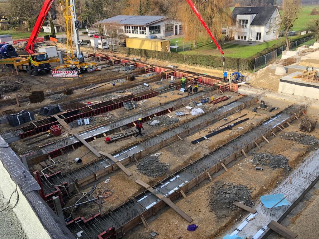 Bauplatz mit Fundamentarbeiten und Kranausrüstung, umgeben von Wohngebäuden und Baustellenmaterial.
