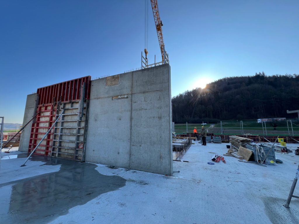 Baustelle mit einer frisch gegossenen Betonwand und einem Kran, der im Hintergrund gegen den Sonnenaufgang arbeitet.