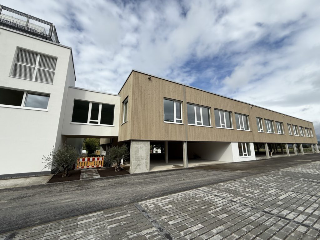 Moderne Gebäude mit Holzverkleidung und großen Fenstern, umgeben von einer gepflasterten Fläche und einem klaren Himmel.