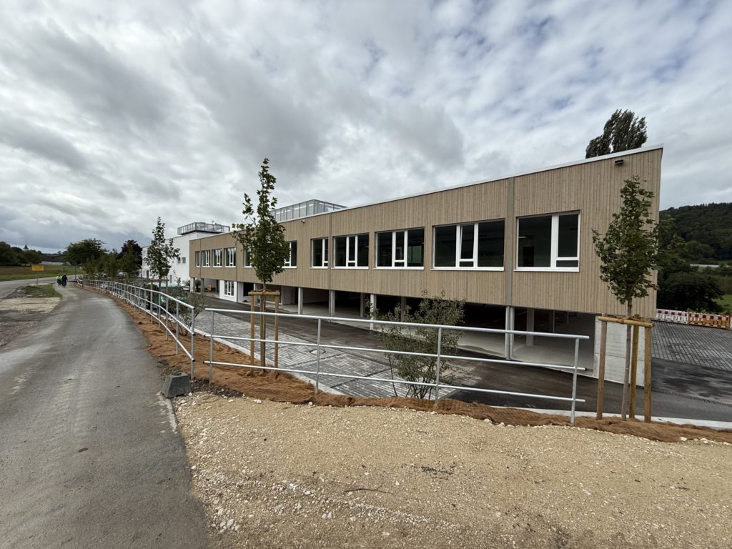 Moderne Schulgebäude mit großen Fenstern, umgeben von neu angelegten Wegen und Bäumen.