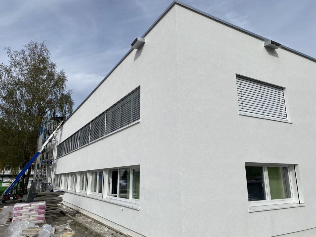 Moderne, weiße Gebäudeaußenansicht mit Fenster und Jalousien, Bauarbeiten im Hintergrund.