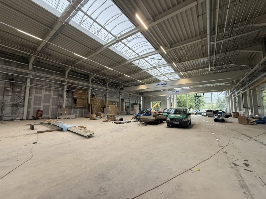 Blick in eine große, leere Halle mit Baustellenmaterial und mehreren Fahrzeugen, die auf dem Boden stehen.