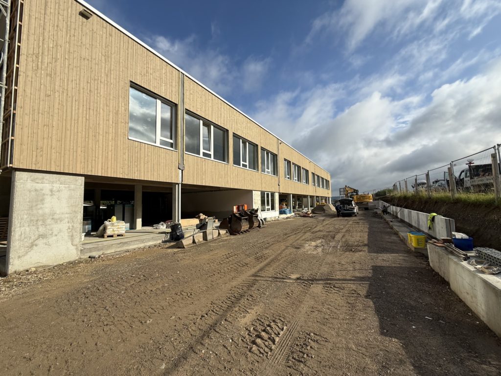 Baufortschritt eines modernen Gebäudes mit Holzfassade und unbefestigtem Weg, umgeben von Baustellenmaterialien.