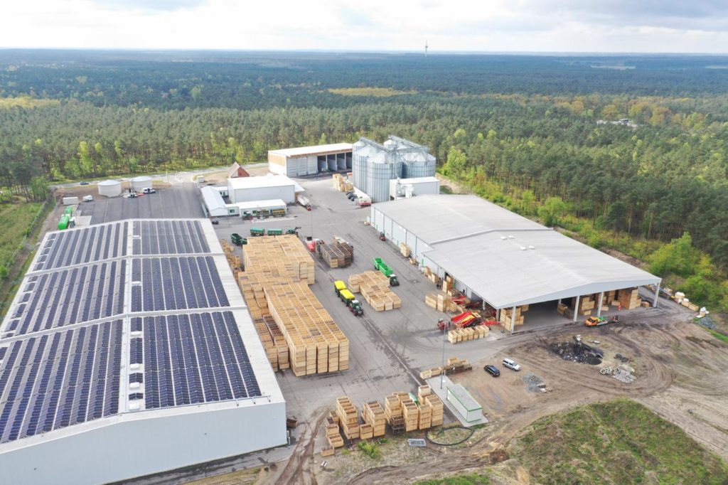 Luftaufnahme eines modernen Werkes mit Solarpanelen und Holzlager in einer bewaldeten Umgebung.