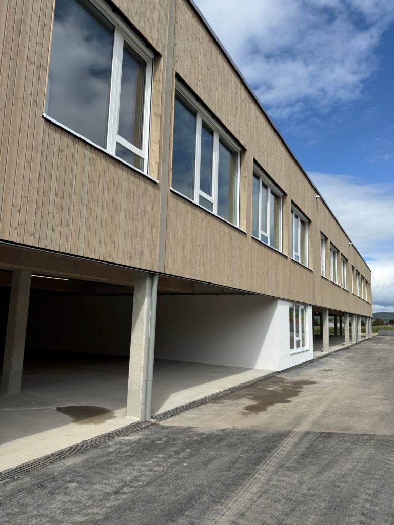 Moderne Gebäudeansicht mit Holzverkleidung und großen Fenstern, darunter ein offener Parkplatz.
